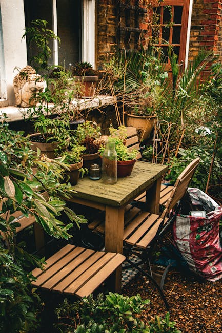 A cozy urban garden patio featuring a weathered wooden table surrounded by matching wooden and metal chairs. The table is adorned with an assortment of potted plants, including succulents and leafy gr