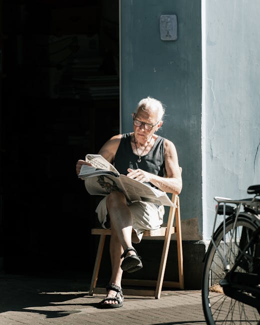 An elderly woman with short gray hair and glasses is seated on a wooden chair outside, reading a newspaper. She is wearing a sleeveless black top, beige shorts, and black sandals. The setting appears 